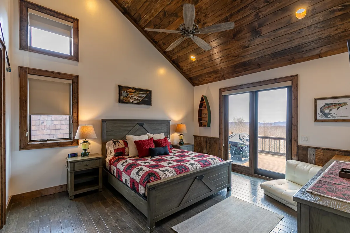This interior image showcases a cozy king bedroom featuring a bed with decorative throw pillows, large windows offering scenic views, a wooden ceiling, and a side table with a lamp. The warm, tranquil ambiance emphasizes comfort and relaxation.