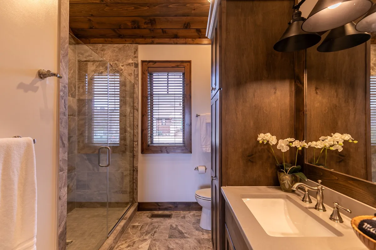 A bright en-suite bathroom with a glass shower enclosure, dual sink vanity, wooden cabinetry, and natural light streaming through the windows. The design is both practical and luxurious, enhancing the suite experience.