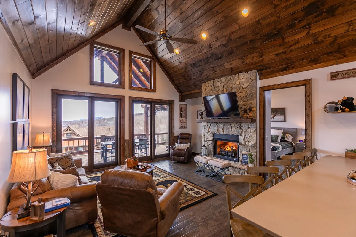 This inviting interior shot showcases a spacious living area with a stone fireplace, large windows framing expansive mountain views, wooden beams, a comfortable seating area, and a dining table nearby. The natural light and open layout highlight both the cozy and communal aspects of the space.