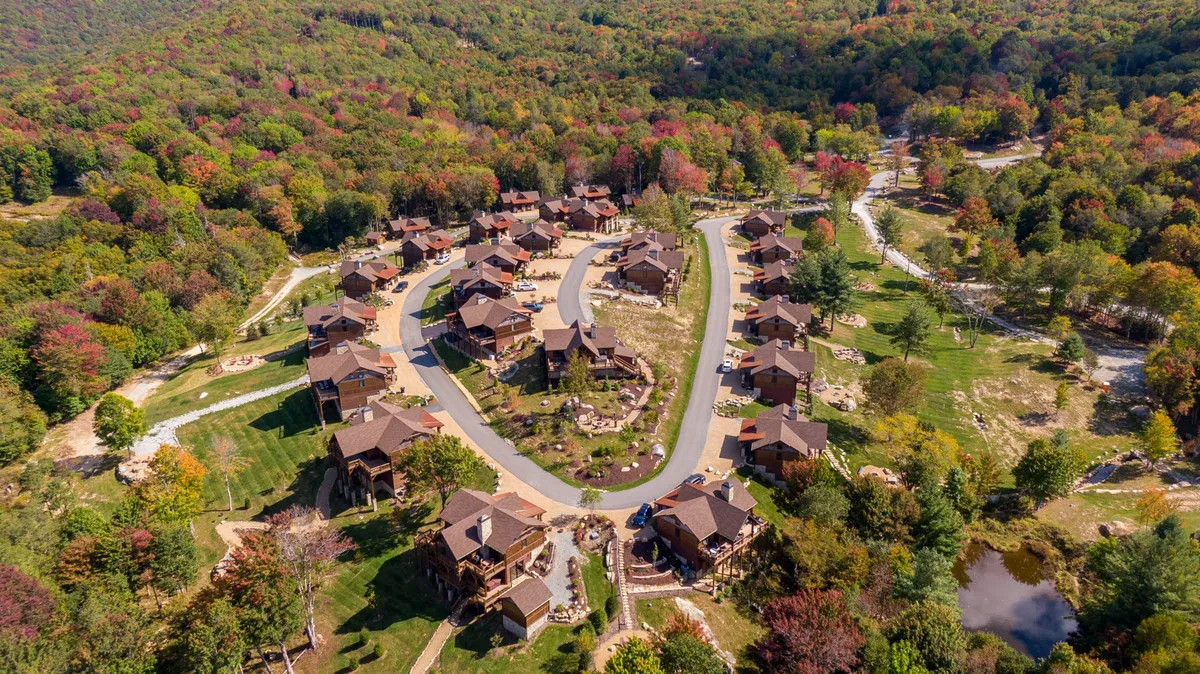 A picturesque exterior scene showing a cluster of cabins nestled among tall trees along a winding road. The autumn setting is alive with warm foliage, and signs of family gatherings, hiking, and outdoor games evoke a sense of exploration and community.