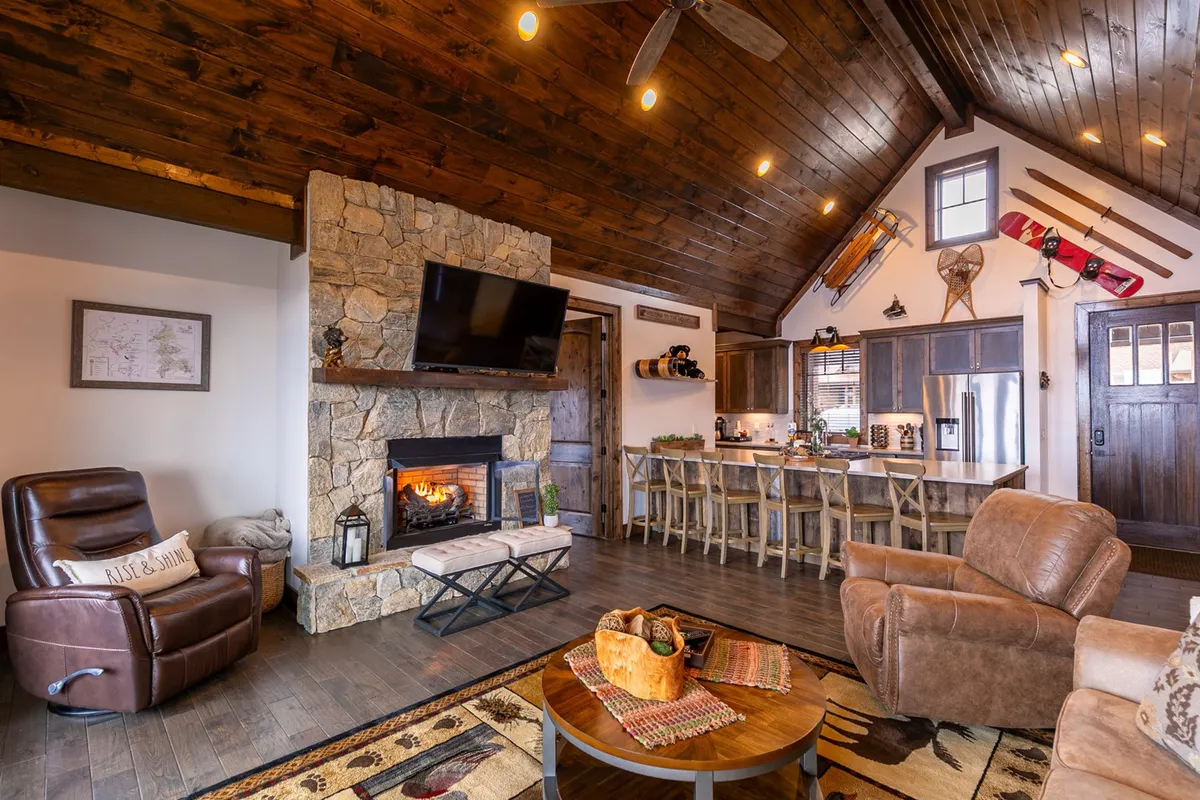 This interior shot offers a wider perspective, featuring a kitchen island adjacent to a welcoming living space with a stone fireplace, wooden ceiling, and leather recliner. The kitchen island is visible, connecting kitchen, dining, and lounging areas—ideal for entertaining and relaxed gatherings.