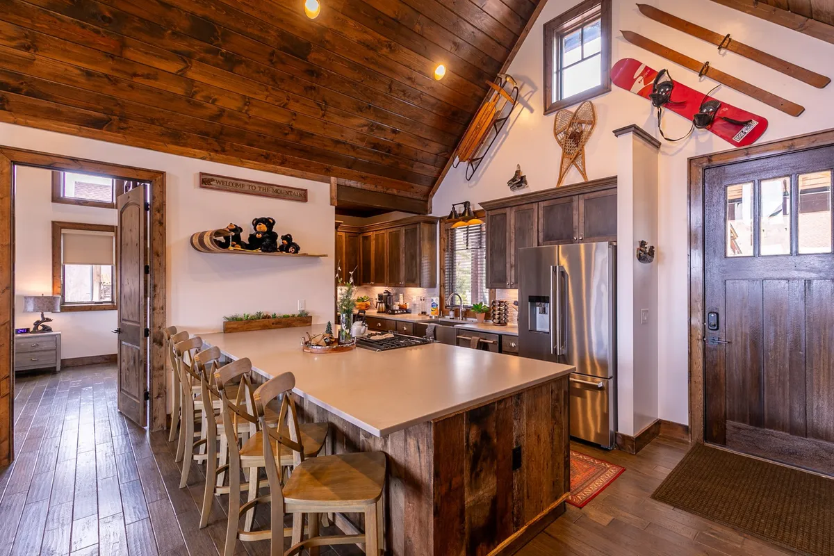 A spacious kitchen with a large island, natural wooden cabinetry, and bar stools provides ample space for meal preparation and socializing. Decorative elements like skis and snowboards add a festive, communal vibe, while the island’s size suggests it easily seats six, ideal for hosting brunches or festive dinners.