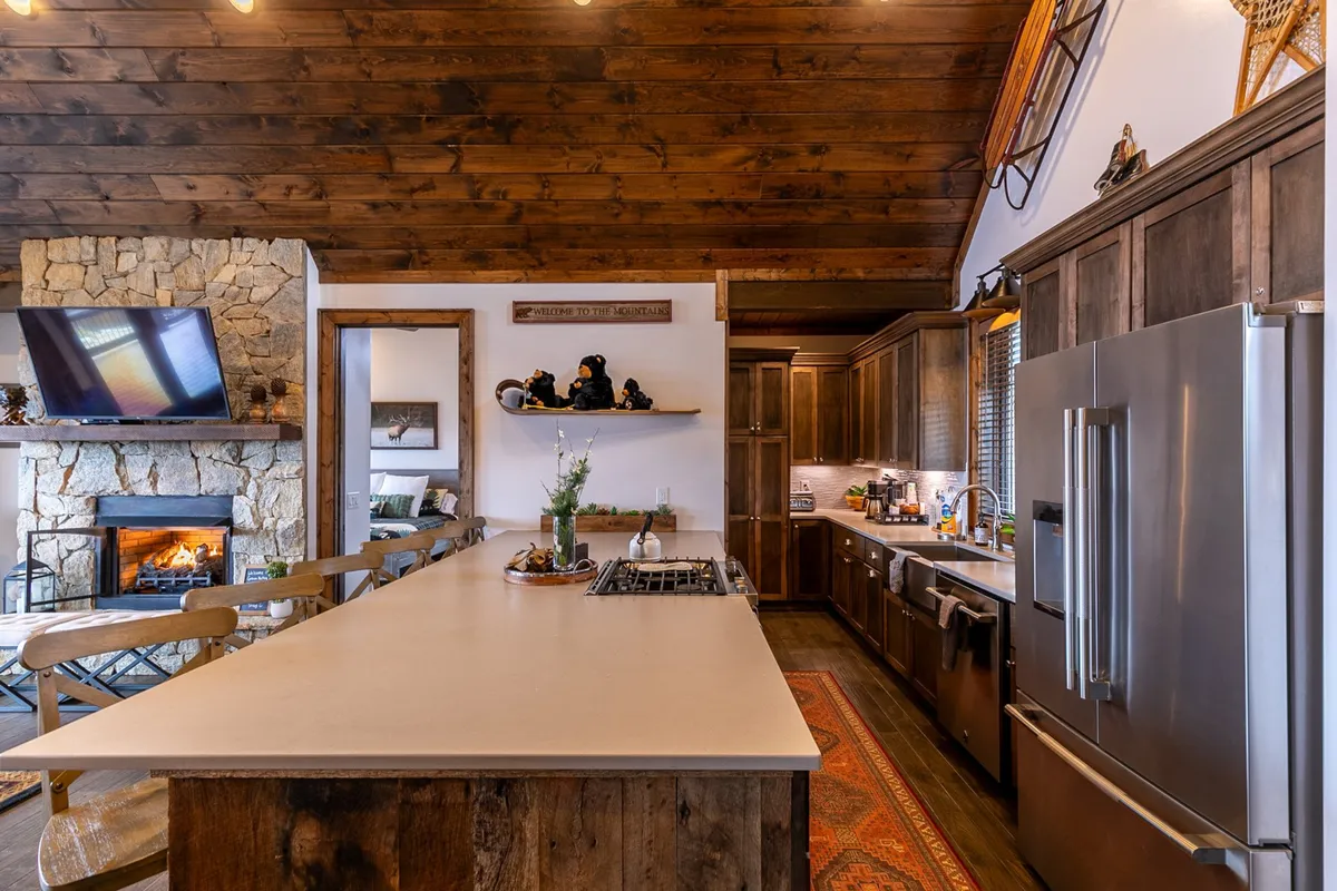 A welcoming view of the kitchen and living area, featuring a large island countertop, modern appliances, a fireplace, and a wooden ceiling. The space is designed for cooking, gathering, and entertaining, with a warm, homey mood.