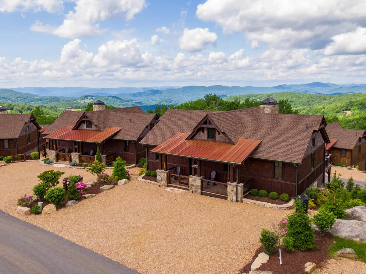 A cluster of wooden cabins nestled amid landscaped gardens, set against a gorgeous mountain backdrop. The scene features tranquil porches where guests are seen relaxing, enjoying coffee, and taking in sweeping long-range views. The soft lighting suggests either early morning or sunset, highlighting the peaceful, romantic vibe.