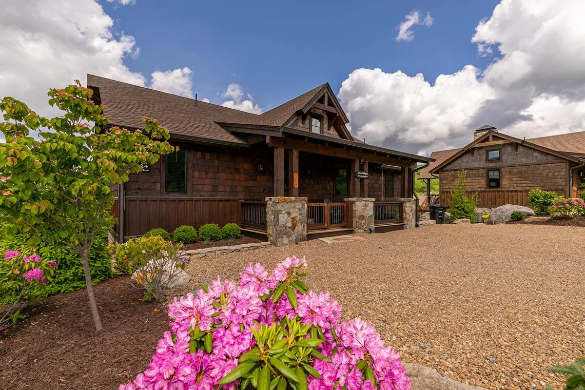 The image features the welcoming exterior of a single-family house with a wooden facade and sturdy stone pillars, surrounded by vibrant flowering plants. The expansive front porch looks out over sweeping mountain views, suggesting the perfect spot for enjoying sunsets, morning coffee, or holiday gatherings in a serene setting.