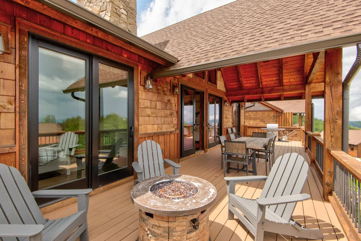 A cozy cluster of Adirondack chairs circles a glowing fire pit on a wooden deck. The surrounding landscape features scenic mountain views and lush greenery, inviting guests to relax, socialize, and enjoy evenings outdoors after a day of adventure.