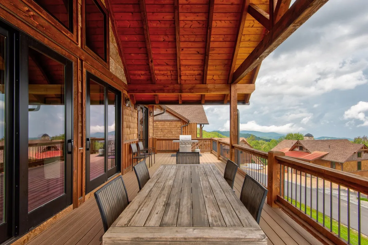 A spacious wooden deck featuring a large table surrounded by chairs, set against a breathtaking backdrop of mountains and expansive views. The deck is adjacent to large windows, seamlessly blending indoor comfort with outdoor living. The setting suggests gatherings, sunset dinners, and communal enjoyment of nature.