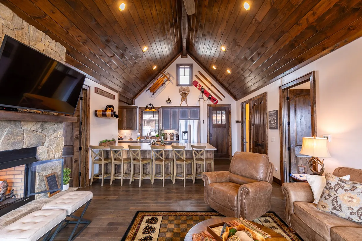 This interior photo features a comfortable living space with a prominent stone fireplace, kitchen island, exposed wooden beams, and plush seating. The scene suggests a lively hub for family gatherings, cooking, and relaxing together.