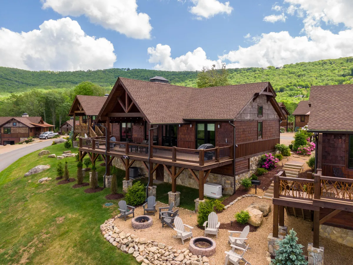 A spacious wooden deck overlooks a lush landscaped garden with a fire pit area in the foreground. Adirondack chairs are arranged around the fire pit, perfect for gathering and relaxation, while the mountain backdrop completes the scene. The setting is inviting for both people and their dogs to enjoy outdoor time together in a safe, comfortable environment.